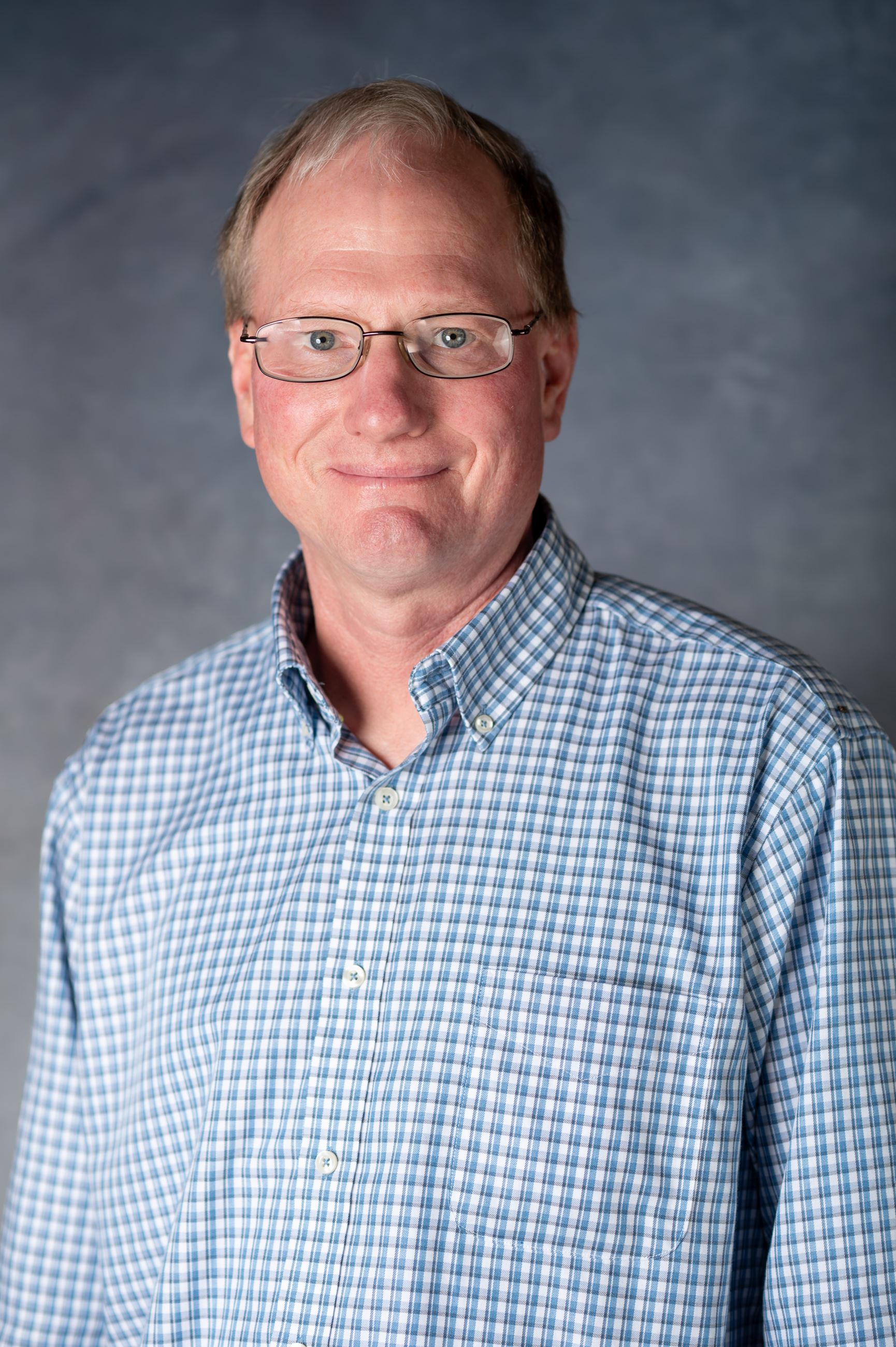 Older man wearing a blue checkered button down and silver wire framed glasses. 