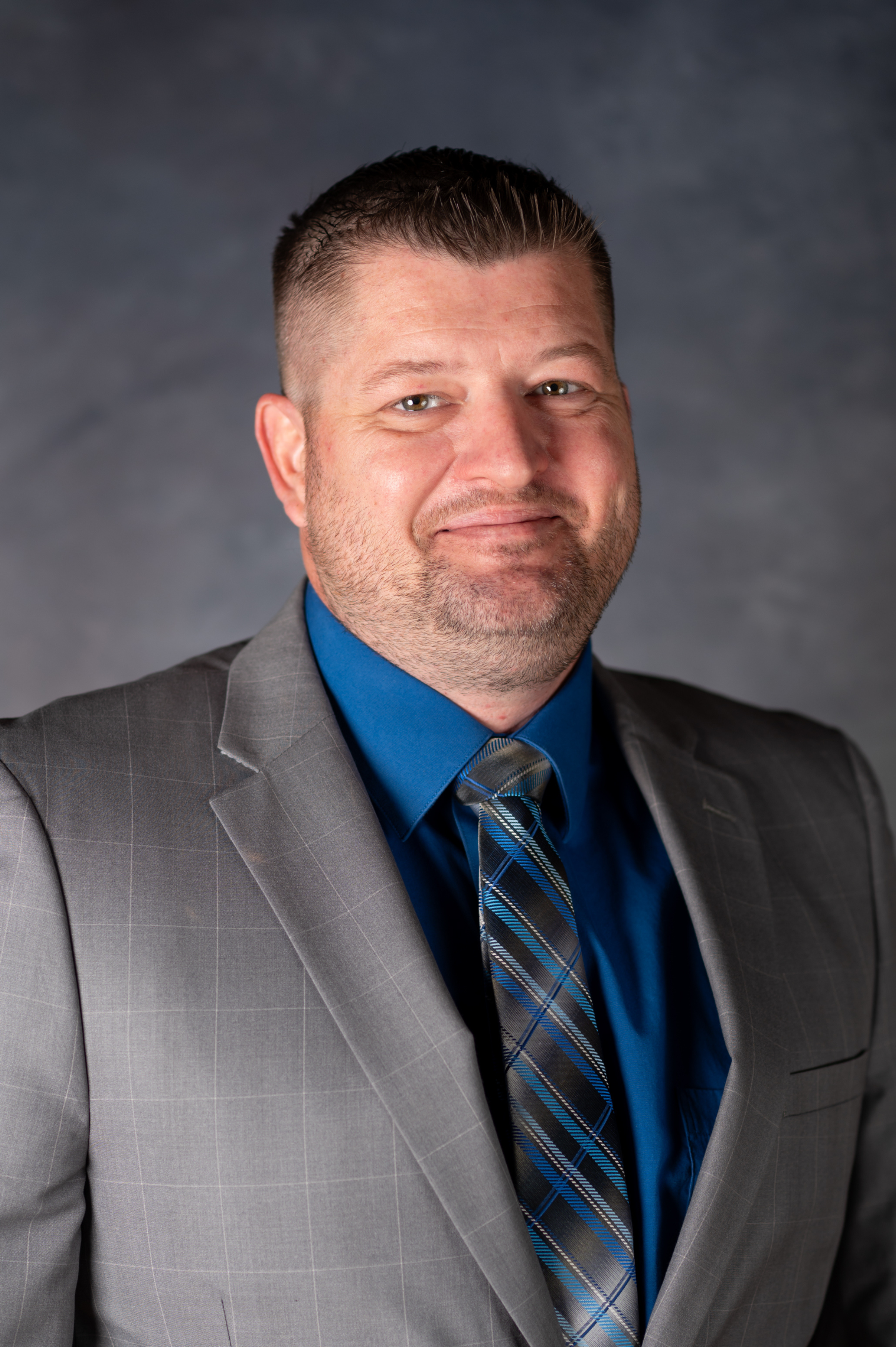 Man with brown hair and slight facial hair smiling at camera in blue shirt and gray suit.