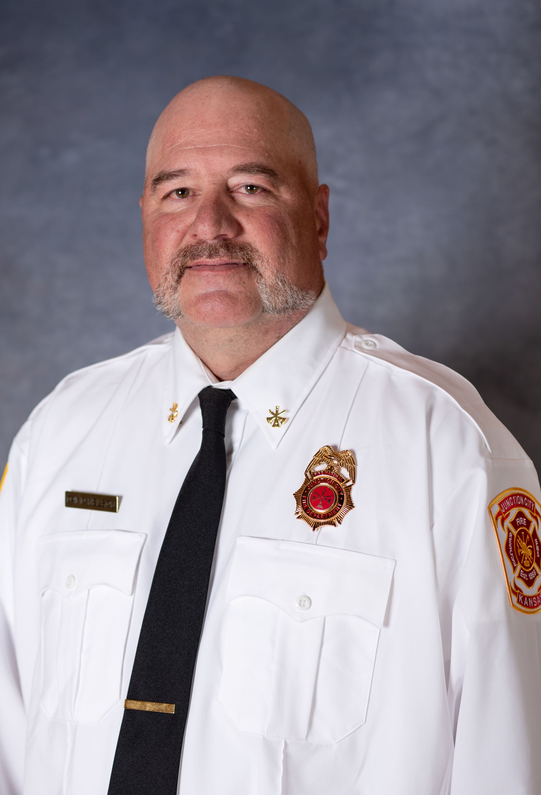 bald man with mustache wearing fireman's dress uniform smiles at camera