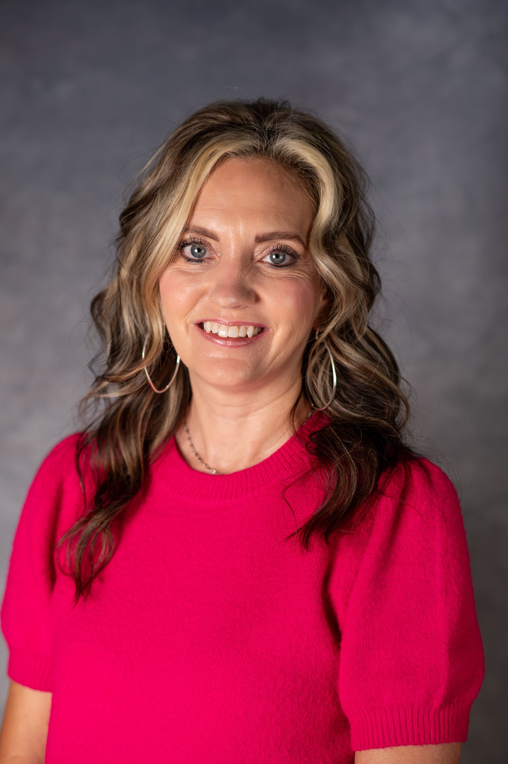 Woman with dirty blonde curly hair smiles for picture in bright pink top.