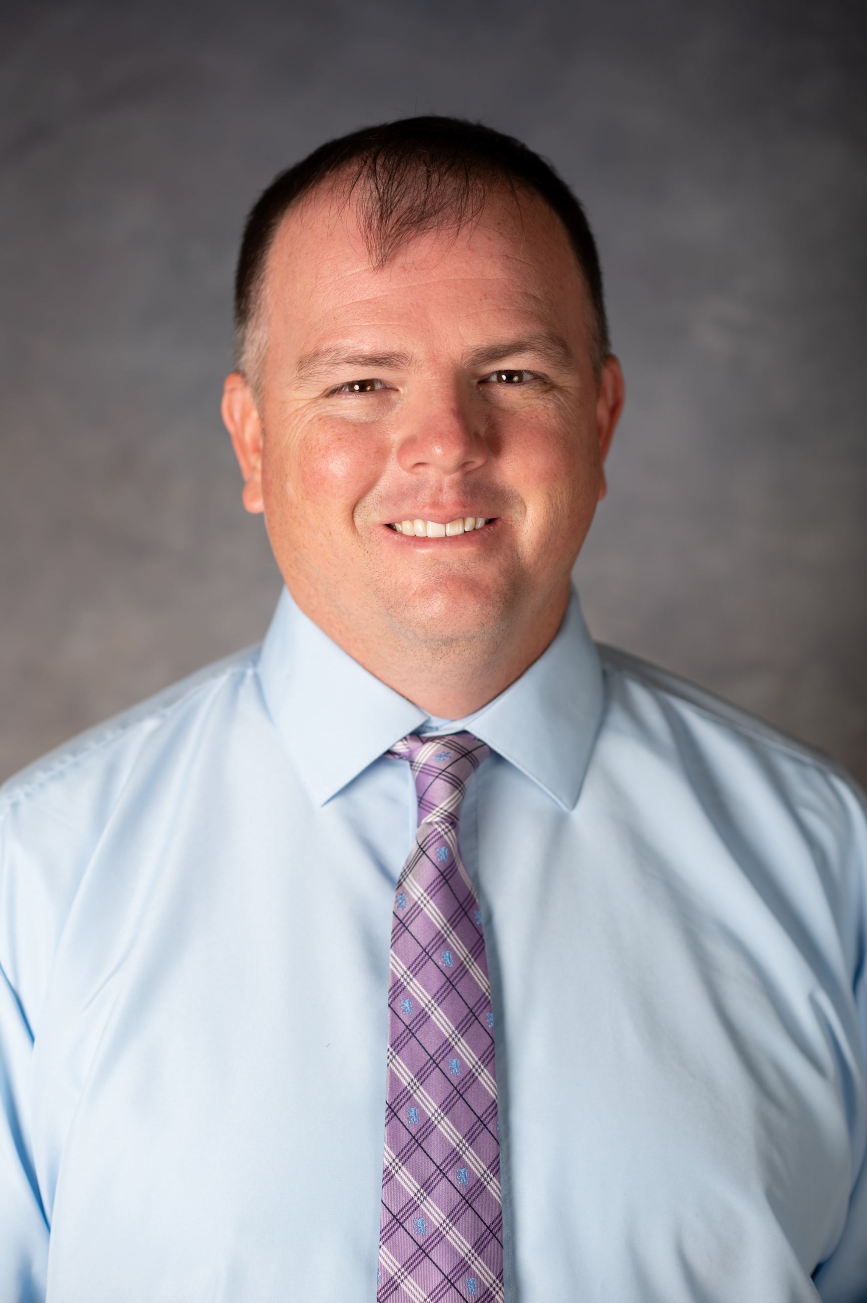 Black with short brown hair smiles for camera wearing blue button down and purple tie