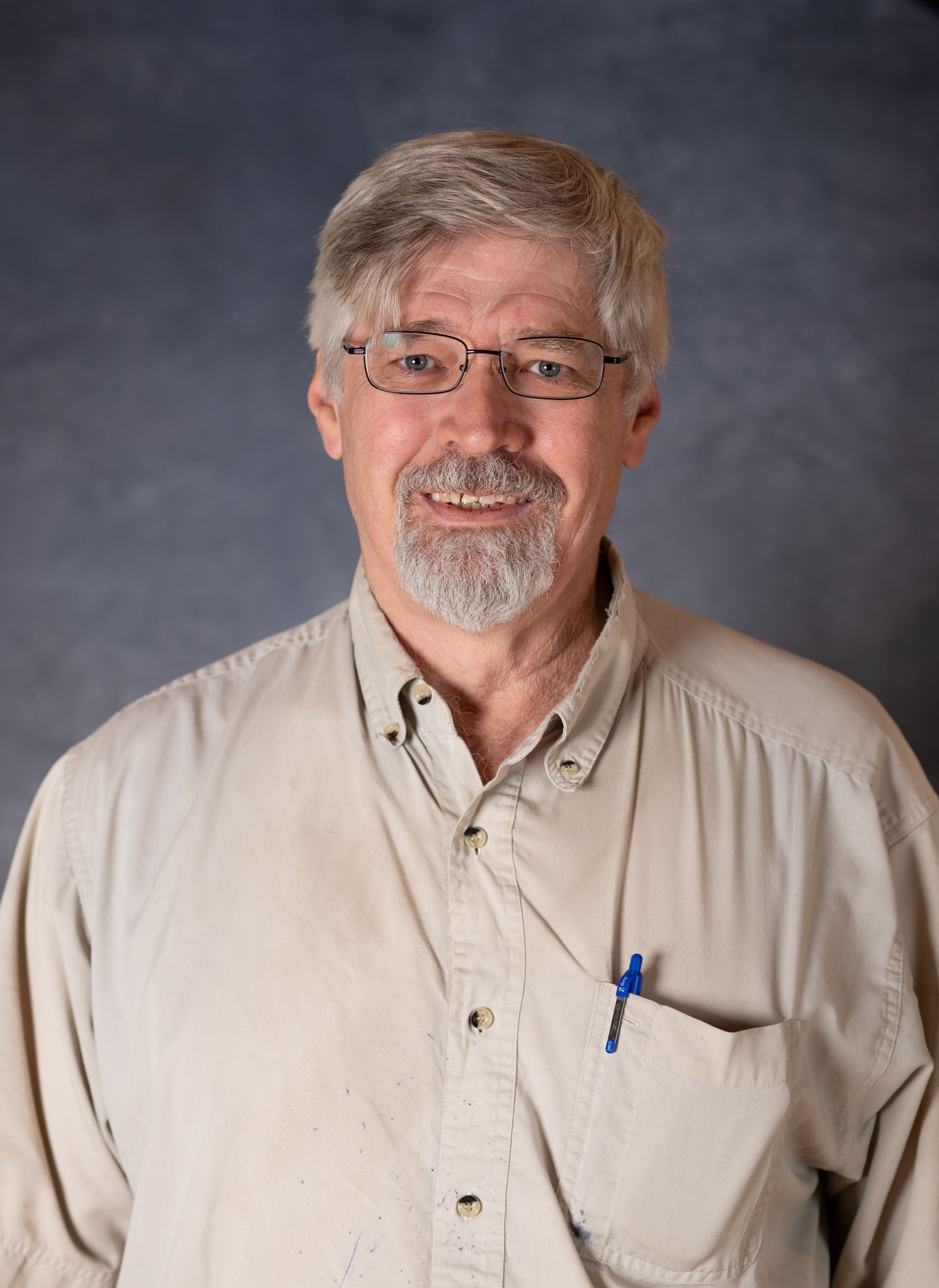 Man with salt and pepper hair and beard wearing tan shirt smiles for camera in tan shirt